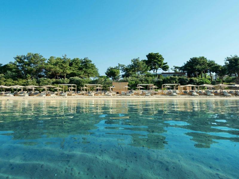 Spiaggia tranquilla con acqua limpida, sdraio e alberi verdi sotto un cielo blu.