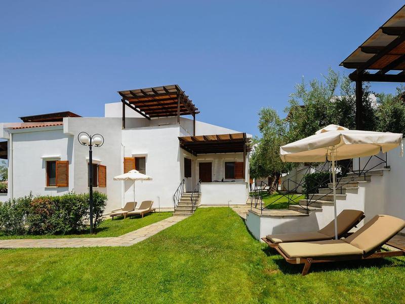 Casa bianca con prato verde, sdraio e ombrellone sotto un cielo azzurro.