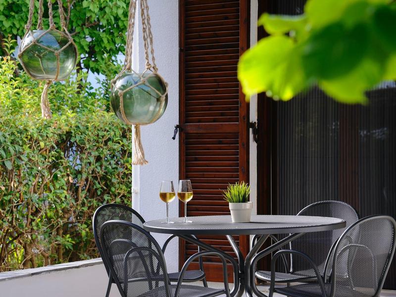 Terrazza con tavolo, sedie e due bicchieri di vino in un ambiente verde.