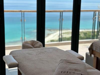 Pokój zabiegowy z łóżkiem do masażu przed balkonem z widokiem na morze i niebieskie niebo.