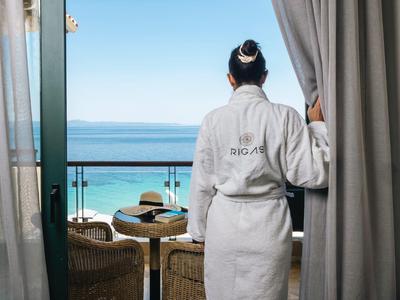 Person im weißen Bademantel steht auf Balkon und blickt aufs blaue Meer unter klarem Himmel.