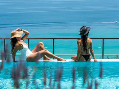 Zwei Frauen entspannen in Badekleidung am Pool mit Blick auf das blaue Meer.