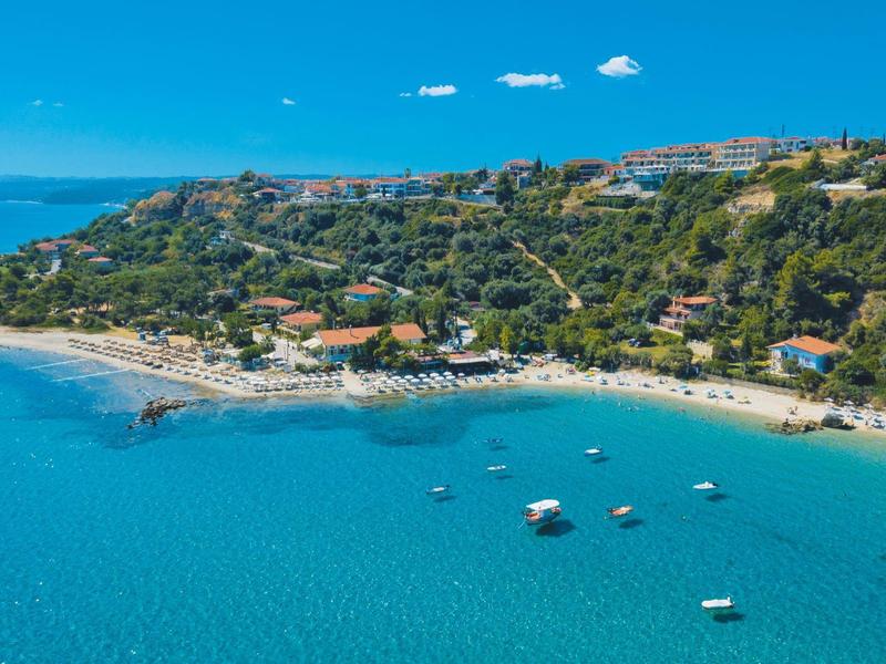 Plaża z czystą niebieską wodą, otoczona zielonymi wzgórzami i budynkami.