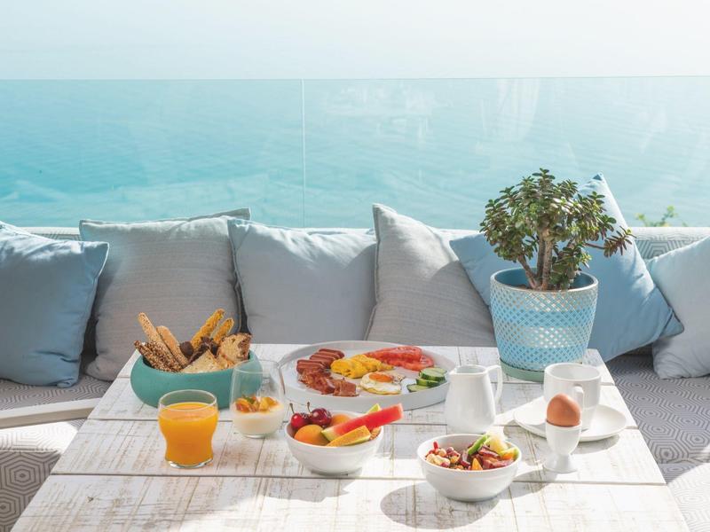 Jasno nakryty stół śniadaniowy z owocami, pieczywem i sokiem na tarasie z widokiem na morze.