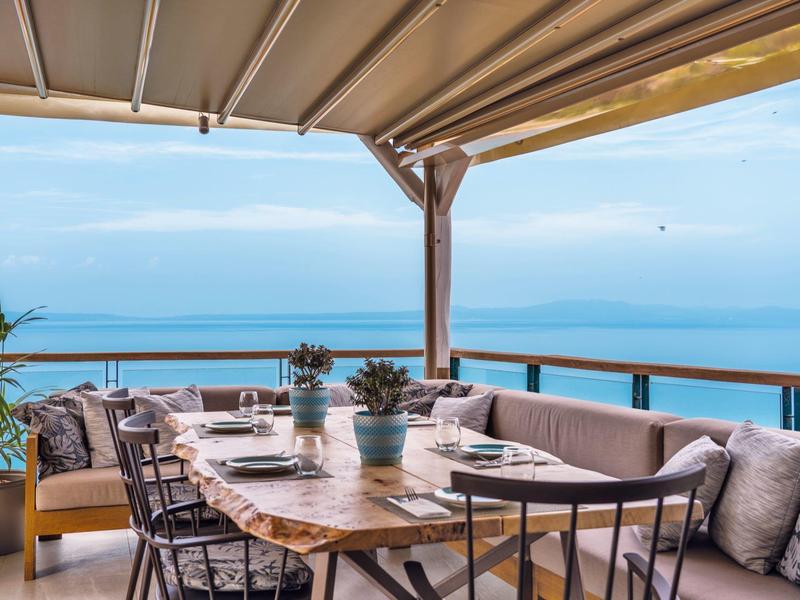 Zadaszony taras zewnętrzny z drewnianym stołem, krzesłami i widokiem na morze w słoneczny dzień.