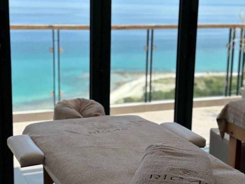 Pokój zabiegowy z łóżkiem do masażu przed balkonem z widokiem na morze i niebieskie niebo.