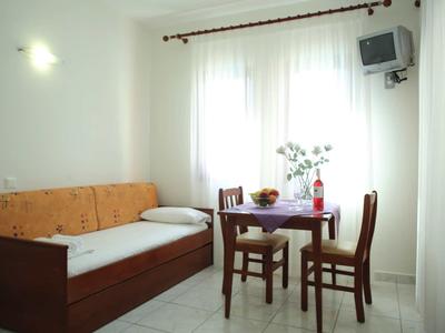 Piccola stanza luminosa con divano, tavolo e due sedie in legno vicino a una finestra con tende.