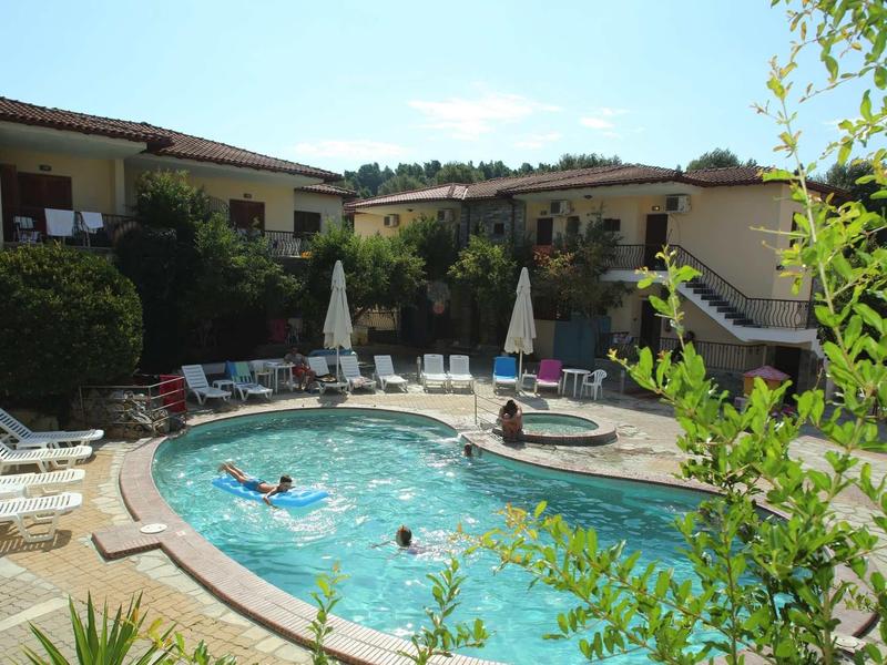 Pool mit mehreren Personen in einem Garten, umgeben von Liegestühlen, Sonnenschirmen und Wohngebäuden.