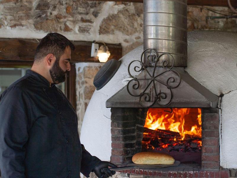 Man die brood bakt in een traditionele stenen oven met open vuur in een rustieke omgeving.
