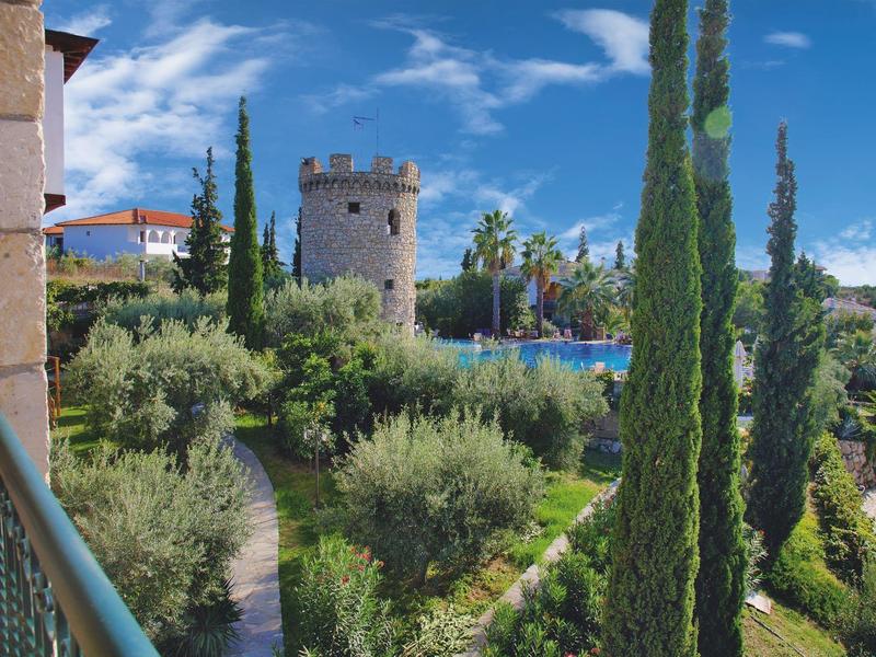 Uitzicht op een tuin met hoge cipressen en een ronde stenen toren op de achtergrond bij heldere hemel.