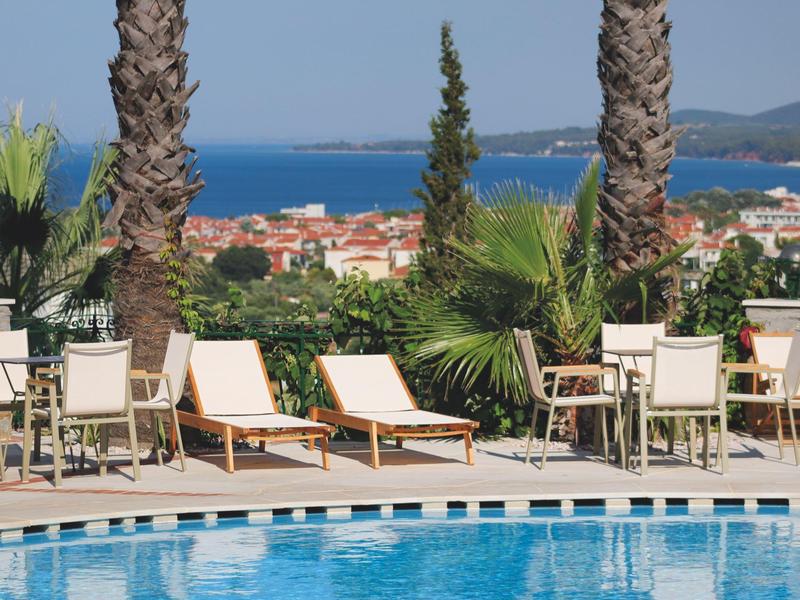 Uitzicht op zwembadgedeelte met ligstoelen en stoelen, palmbomen en zee op de achtergrond.