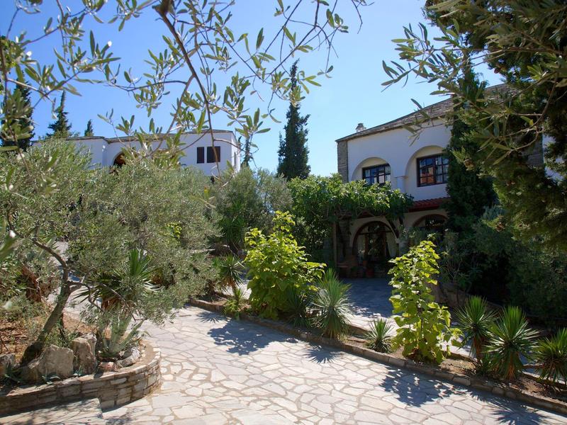 Een geplaveid pad door een tuin met olijfbomen naar een wit huis met blauwe luiken.