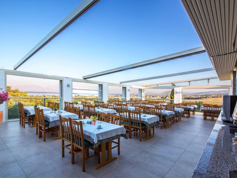 Ruim terras met houten tafels en stoelen met uitzicht op zee onder een blauwe lucht.
