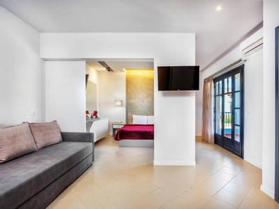 Habitación de hotel luminosa con sofá, televisor de pared, puerta abierta al dormitorio y ventana grande.