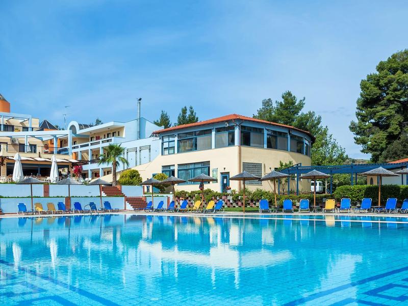 Piscina exterior grande con sombrillas y edificios turísticos alrededor bajo un cielo azul despejado