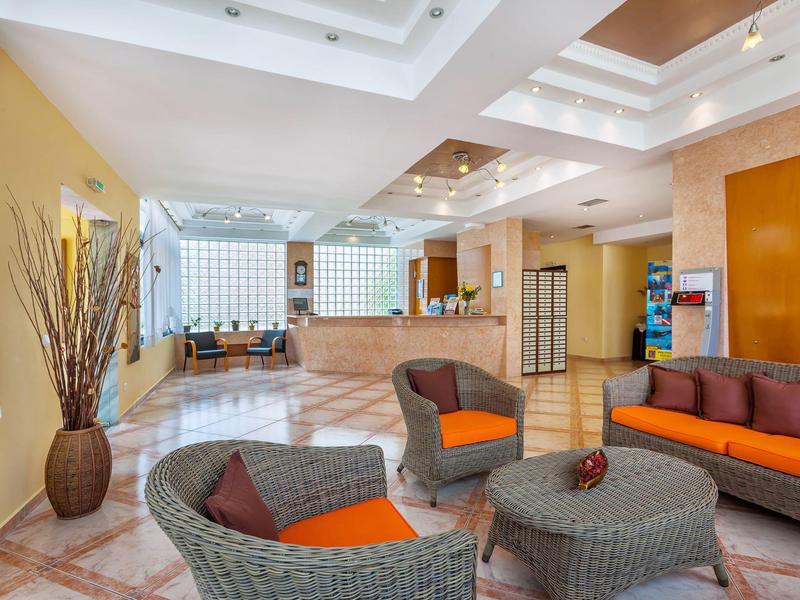 Luminoso vestíbulo de hotel con muebles de mimbre, cojines naranjas y un jarrón alto de flores.