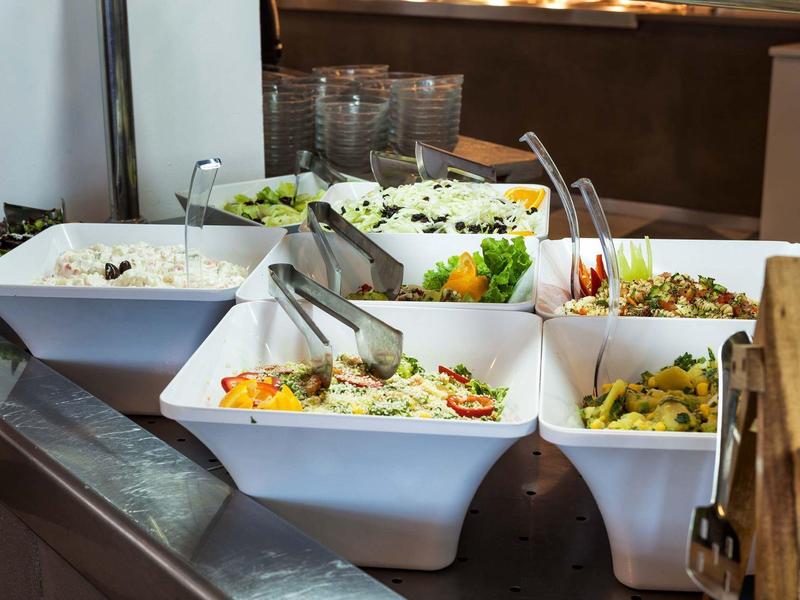 Bufé con diferentes ensaladas en recipientes blancos, servido con pinzas y vasos al fondo.