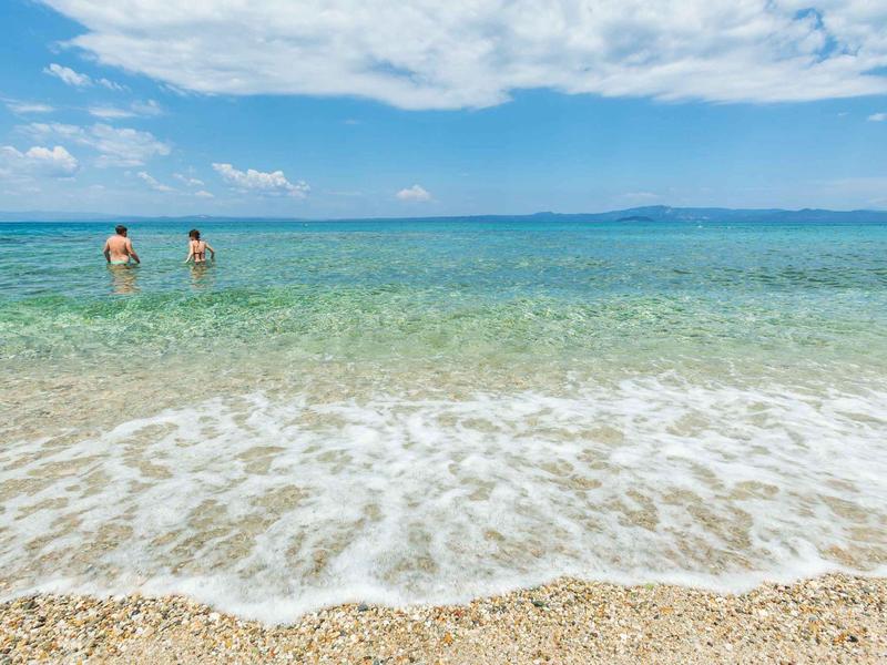 Dos personas están en el agua clara y poco profunda de una playa de arena bajo un cielo azul.