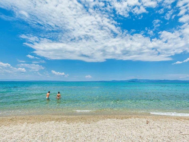 Playa de arena blanca con agua clara y tranquila, y dos personas jugando en el mar bajo un cielo azul.