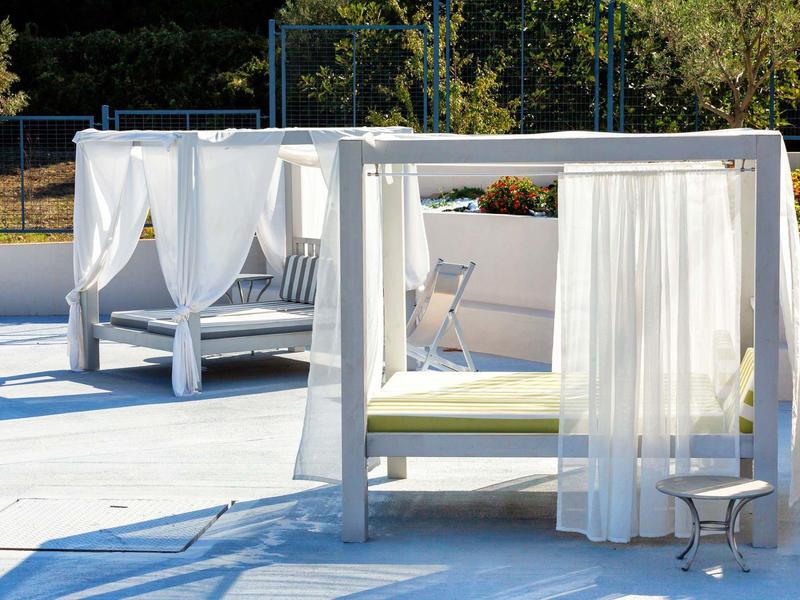Dos camas modernas blancas con cortinas en una terraza con suelo azul, rodeadas de árboles.