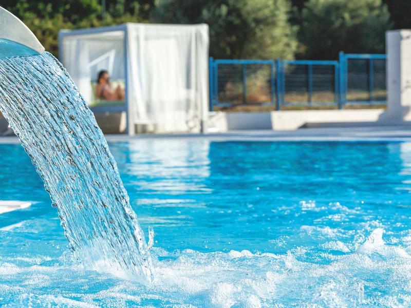 Agua que cae en una piscina azul clara con una cama balinesa y vegetación al fondo.