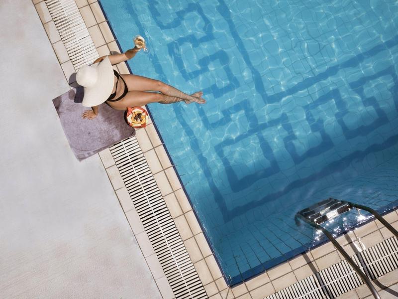 Mujer relajándose en el borde de la piscina sobre una toalla, usa sombrero, piscina azul con patrón y escalera visible