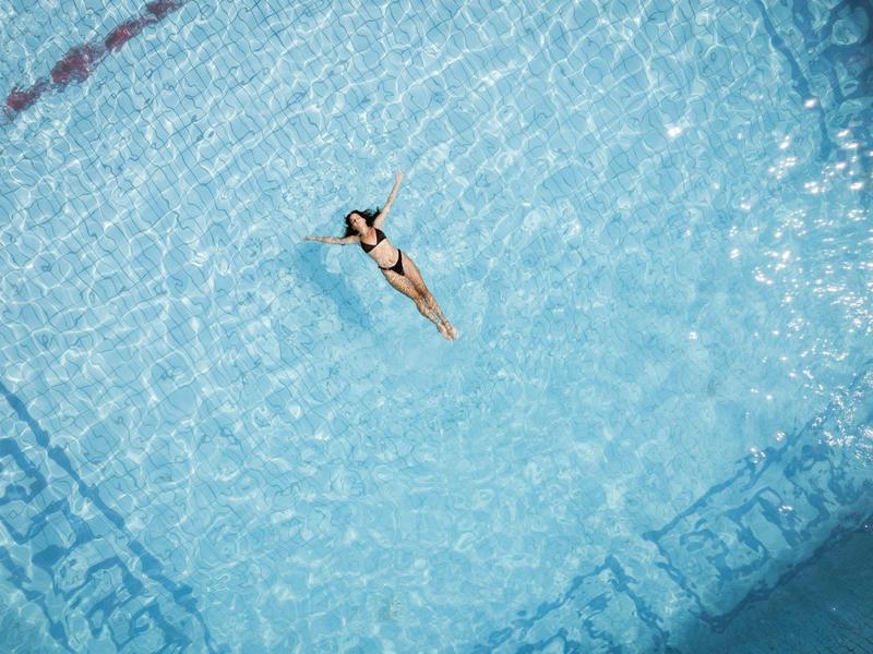 Mujer nadando relajada de espaldas en una gran piscina azul clara.