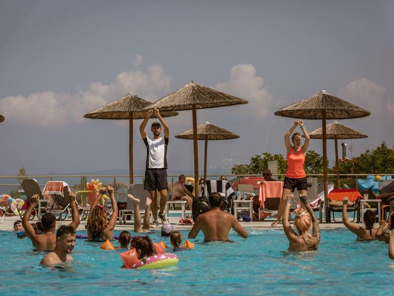 Personas hacen gimnasia acuática en la piscina con instructores al borde y sombrillas de paja.