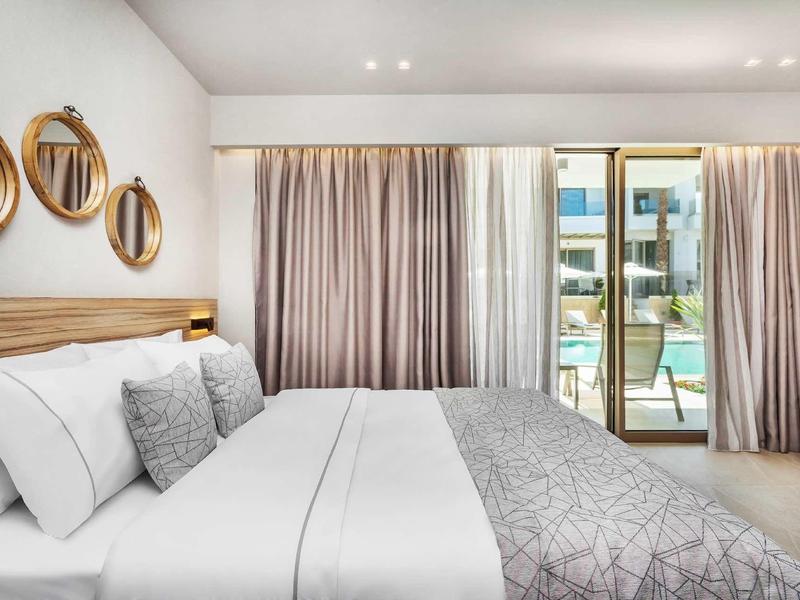 Jasny pokój hotelowy z dużym łóżkiem, zasłonami i tarasem z widokiem na wodę.