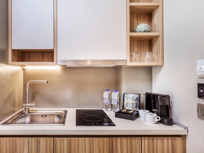 Nowoczesna hotelowa kuchnia z zlewem, płytą grzewczą, ekspresem do kawy i minimalistycznymi drewnianymi szafkami.