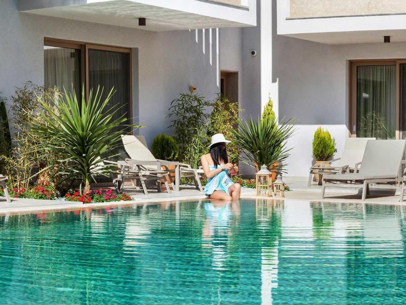 Kobieta siedzi na brzegu basenu w hotelowym ogrodzie z leżakami i roślinami.