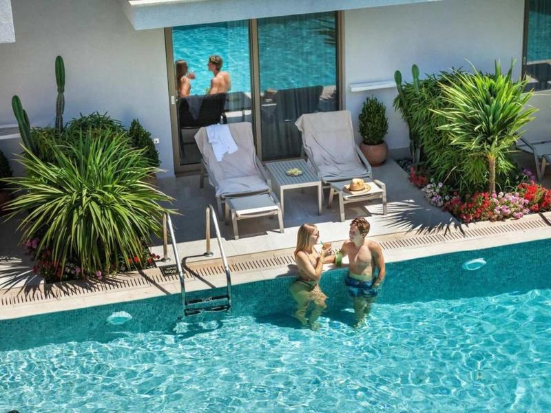 Dwie osoby pijące napój w przejrzystej wodzie basenu obok leżaków i roślin.