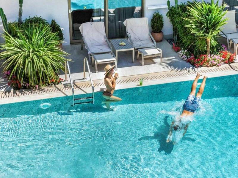 Dwoje dzieci skacze do czystego basenu obok leżaków i zielonych roślin.