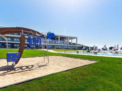 Spielplatz mit Schaukel auf Gras, Sandfläche und modernes Hotelgebäude im sonnigen Freien.