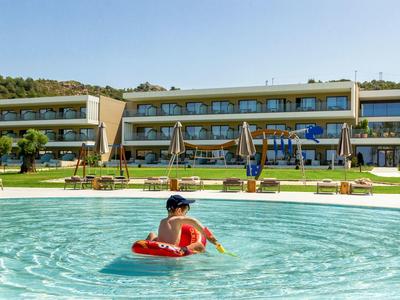 Ein Kind schwimmt auf einem roten Schwimmring in einem großen blauen Pool vor einem Hotelgebäude bei Sonnenschein.