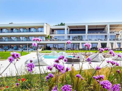 Modernes Hotel mit Schwimmbad, Sonnenliegen und lila Blumen im Vordergrund unter blauem Himmel.