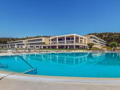 Großer, blauer Hotelpool mit Stufen und Sonnenschirmen vor modernem Gebäude und bewaldetem Hügel.
