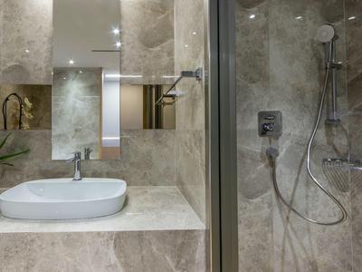 Moderner Badbereich mit grauen Fliesen, weißem Waschbecken, Spiegel und Dusche mit Glasabtrennung.