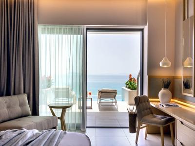 Helles Hotelzimmer mit Bett, Stuhl, Schreibtisch, Glas-Schiebetür und Meerblick auf Balkon.