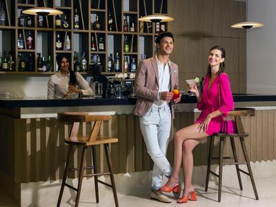 Zwei elegant gekleidete Personen sitzen an einer modernen Bar mit Holzstühlen und Weinregal.