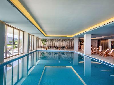 Hallenbad mit klarem Wasser, großen Fenstern und Liegestühlen an der Seite, helle Deckenbeleuchtung.