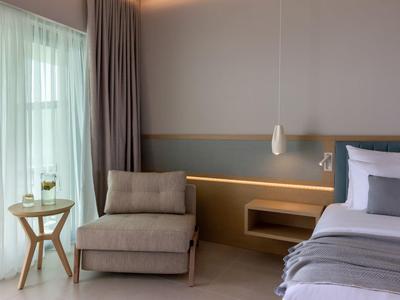 Modernes Schlafzimmer mit grauem Sessel, Holztisch, großem Bett und weißen Vorhängen am Fenster.