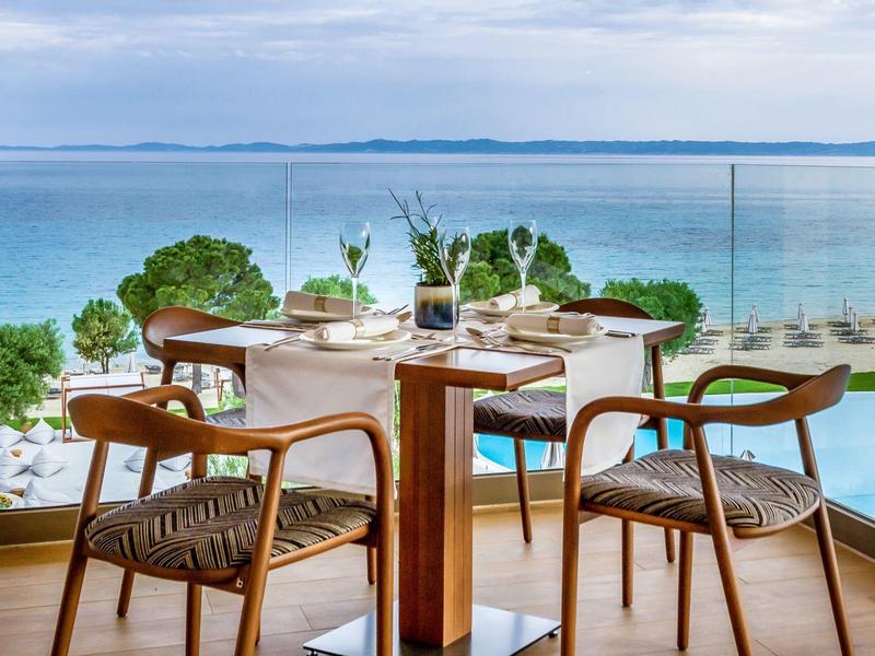 Moderner Balkon mit Holztisch und vier Stühlen, Glasgeländer, Blick auf blaues Meer und Himmel.