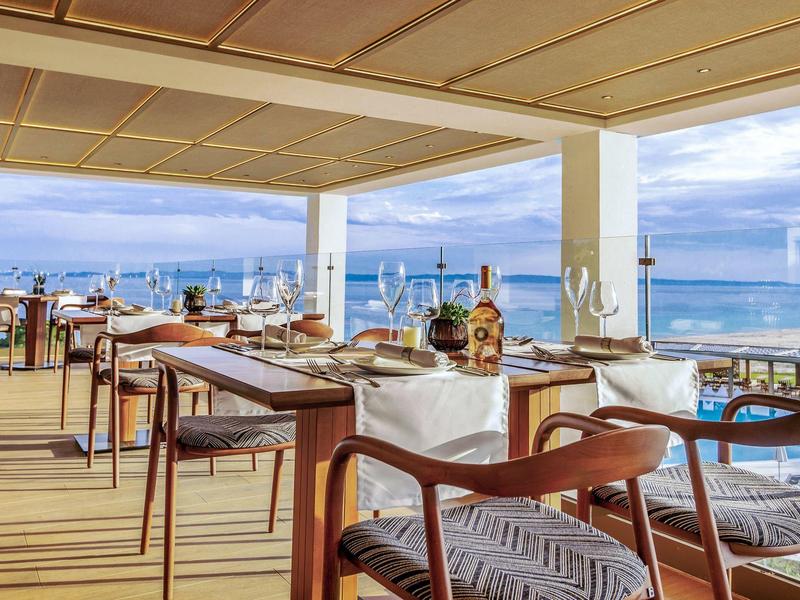 Offenes Restaurant mit Holztischen, Stühlen und Meerblick unter blau-weiß bewölktem Himmel.