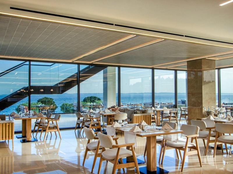 Helle, moderne Cafeteria mit Holz- und weißen Möbeln, großen Fenstern und Blick auf Stadt und Meer.