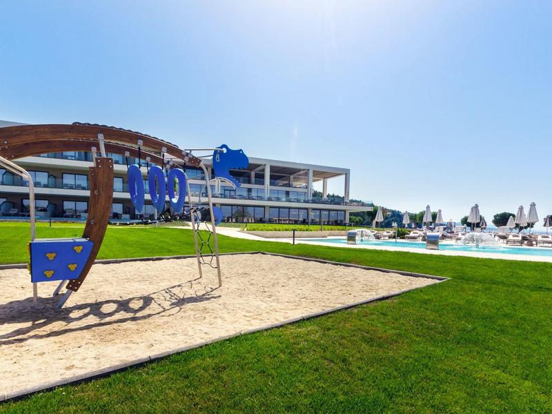 Spielplatz mit Schaukel auf Gras, Sandfläche und modernes Hotelgebäude im sonnigen Freien.