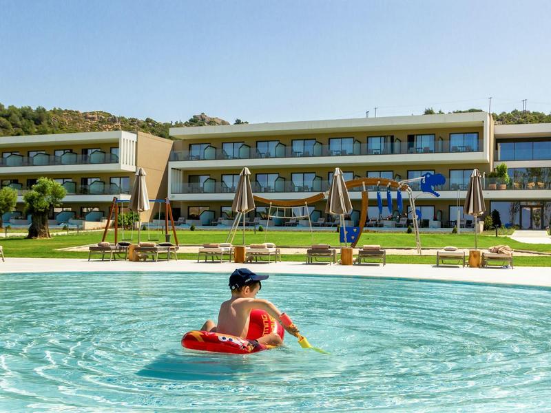 Ein Kind schwimmt auf einem roten Schwimmring in einem großen blauen Pool vor einem Hotelgebäude bei Sonnenschein.