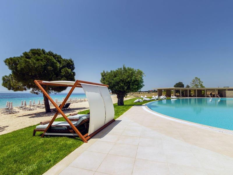 Pool mit blauer Wasserfläche, Liegen aus Holz und weißen Stoffen, Rasen und Strand mit Sonnenschirmen.