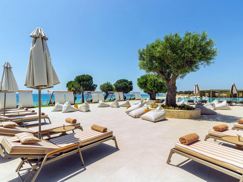 Strandresort mit Liegen, Sonnenschirmen, Olivenbäumen und Blick auf das blaue Meer.