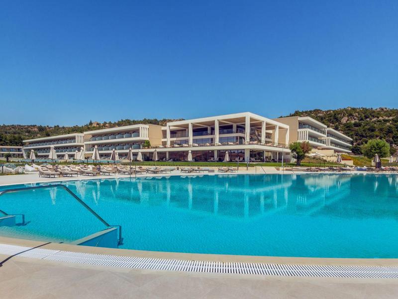 Großer, blauer Hotelpool mit Stufen und Sonnenschirmen vor modernem Gebäude und bewaldetem Hügel.